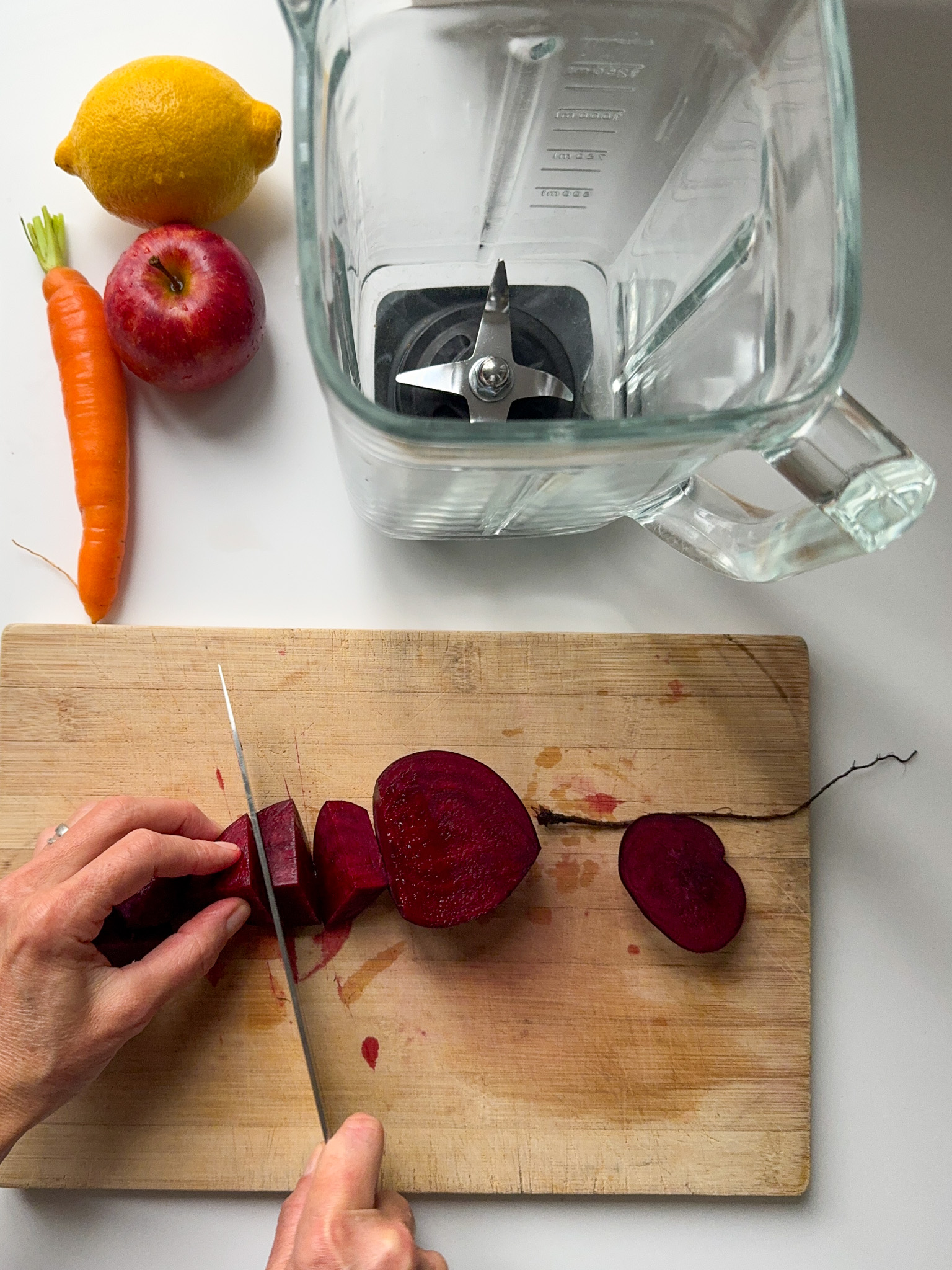 Cutting beetroot on a wood board with a Apple, Carrot and Lemon on the side of a blender.