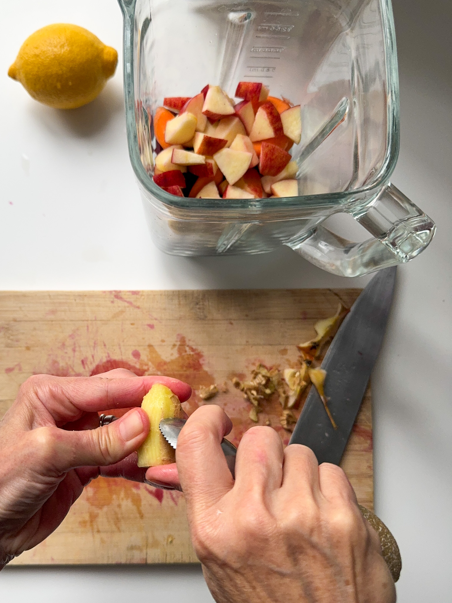 Peeling a piece of ginger with a tea spoon, a lemon next to a blender with cut beetroot and apple