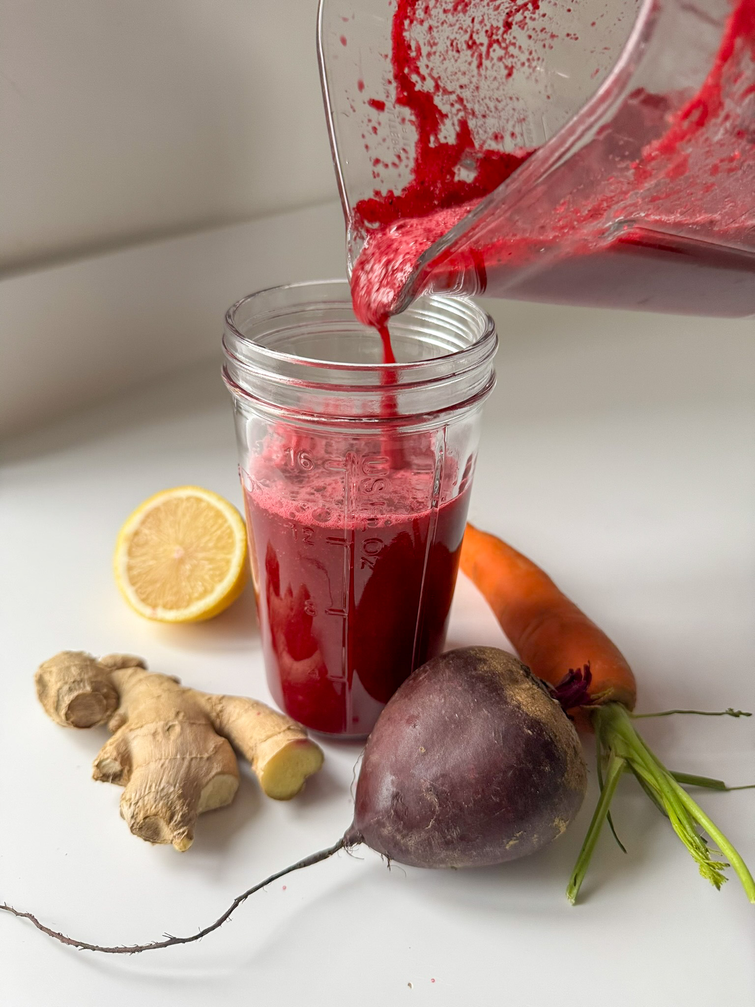 Pouring Beetroot and Ginger Juice into a glass surround by beetroot, lemon, carrot and ginger.