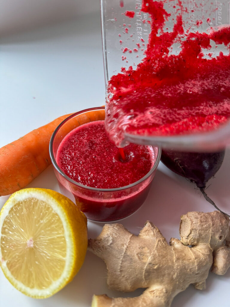 Pouring beetroot juice on a glass. Surround by fresh Beets, Ginger, Lemon and carrots