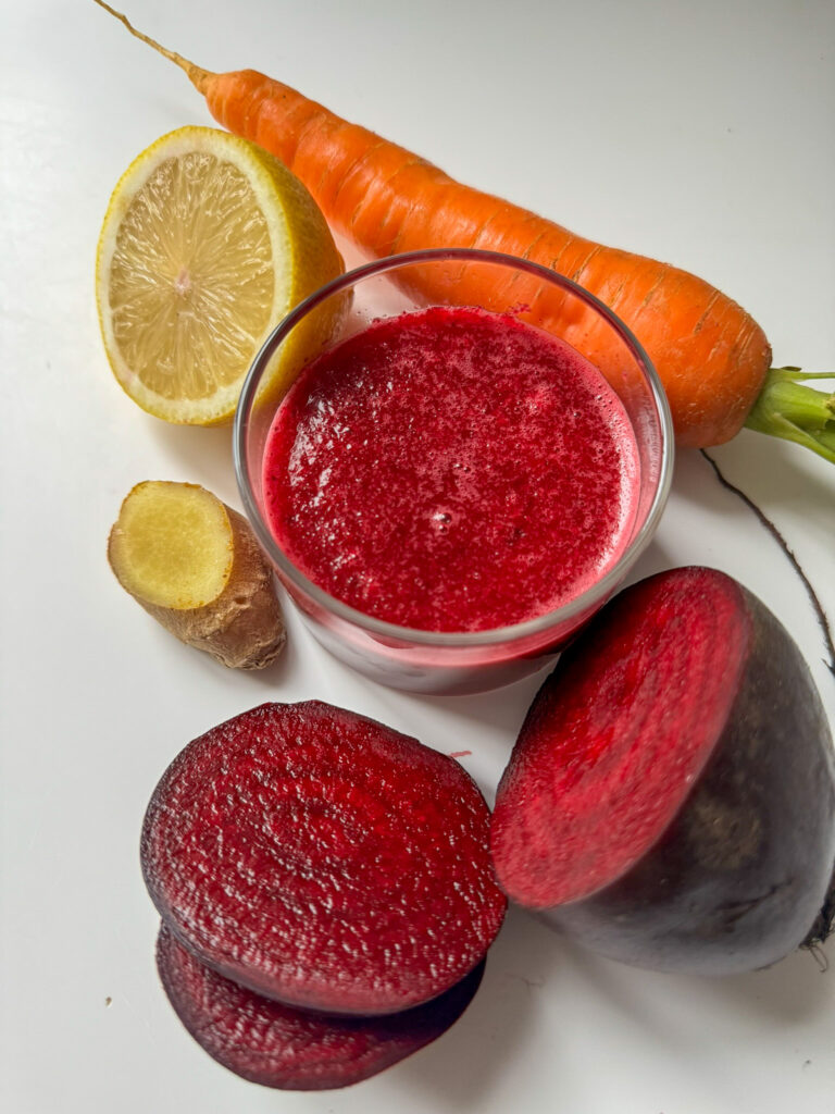 Beetroot, Ginger, Carrot and Apple Juice in a glass surround by Beetroot, Carrot, lemon and Ginger from top.