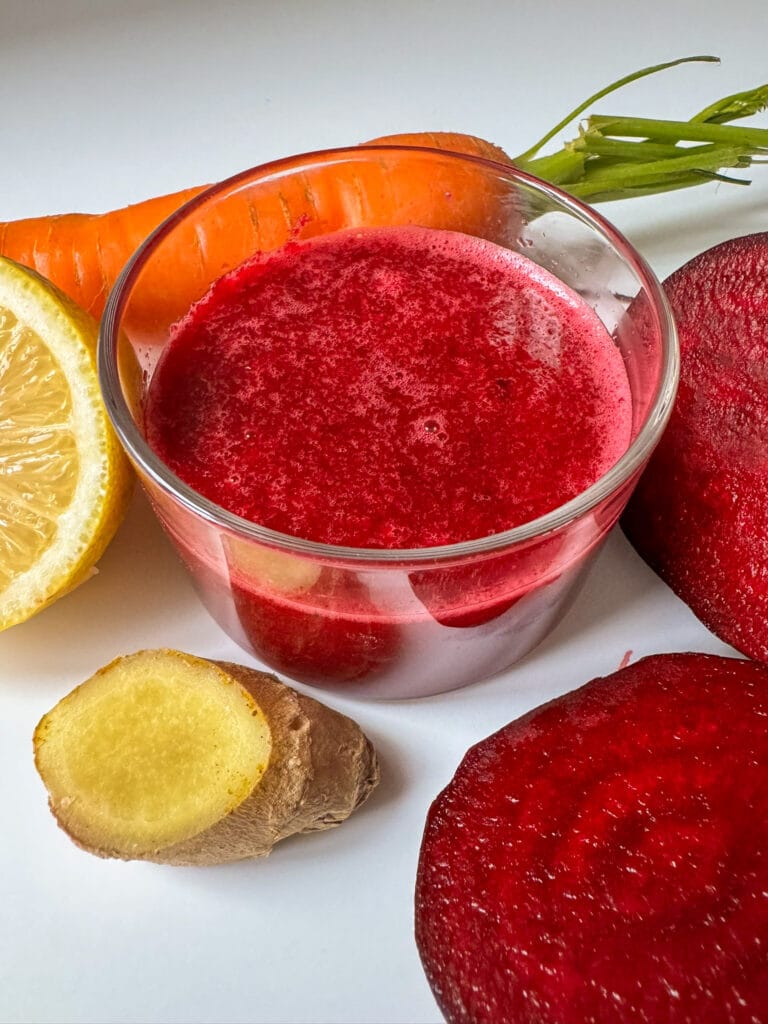 Beetroot, Ginger, Carrot and Apple Juice in a glass surround by Beetroot, Carrot, lemon and Ginger