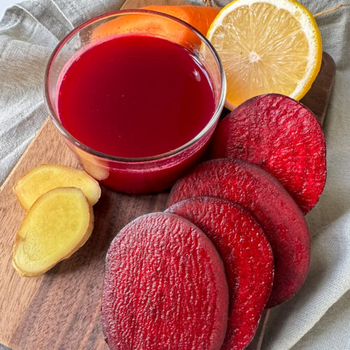 Beetroot, Ginger, Carrot and Apple Juice in a glass surround by Beetroot, Carrot, lemon and Ginger on a wood board