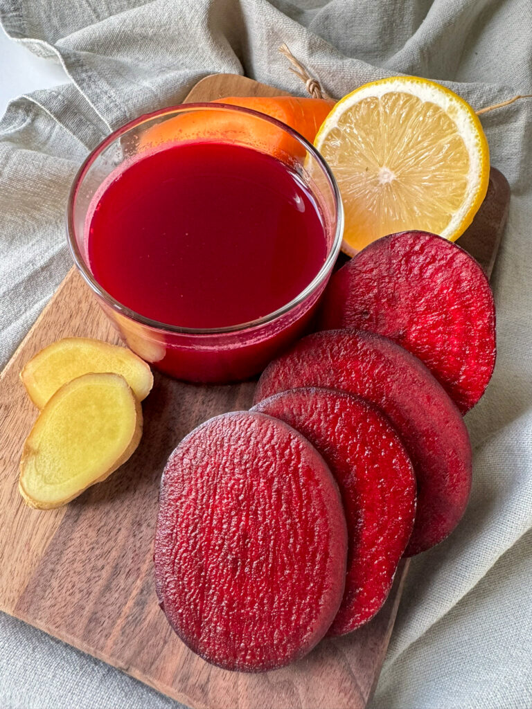 Beetroot, Ginger, Carrot and Apple Juice in a glass surround by Beetroot, Carrot, lemon and Ginger on a wood board