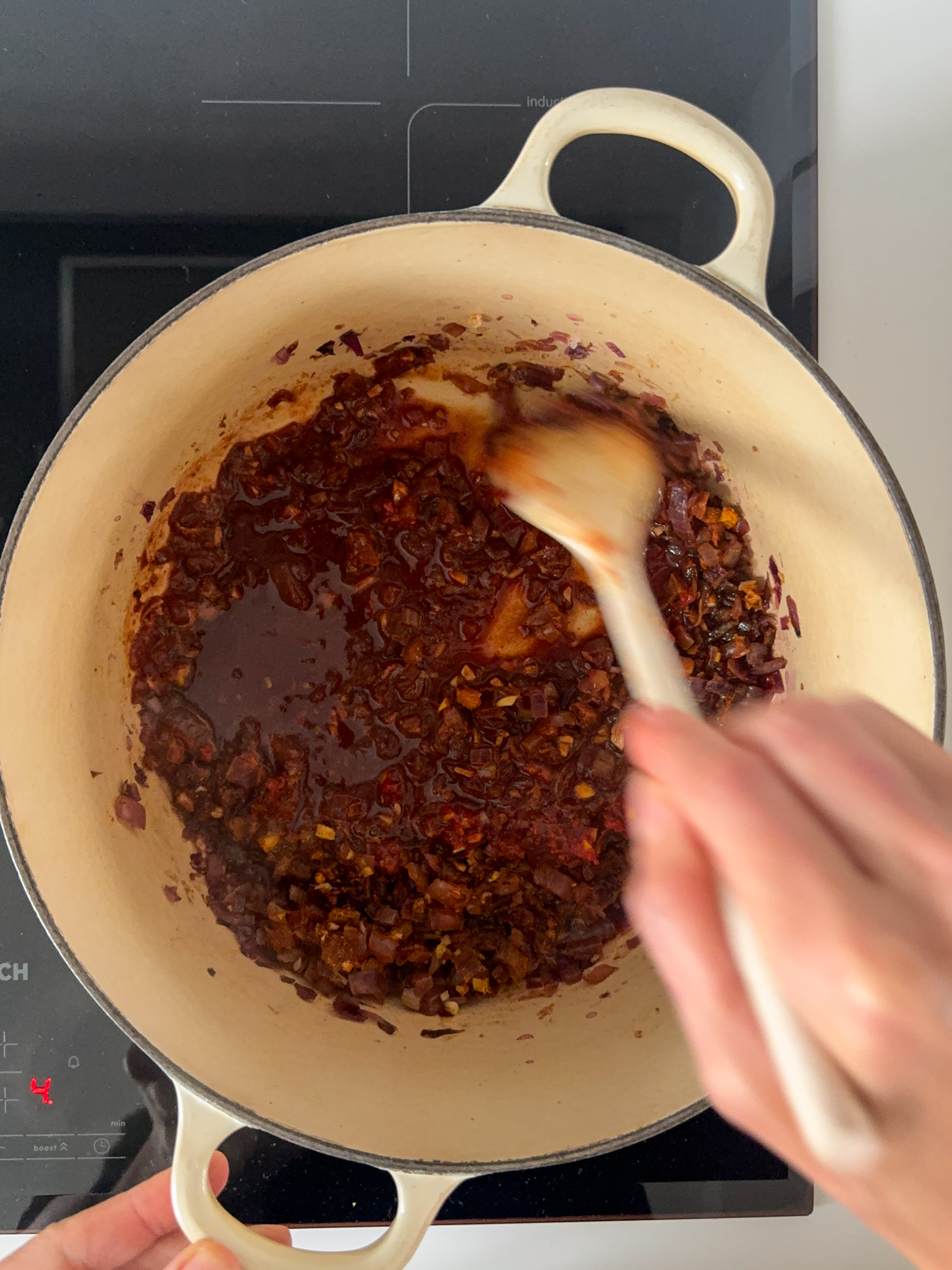 A white dutch oven with red onion, garlic, spices and red wine being stir with a wood spoon.