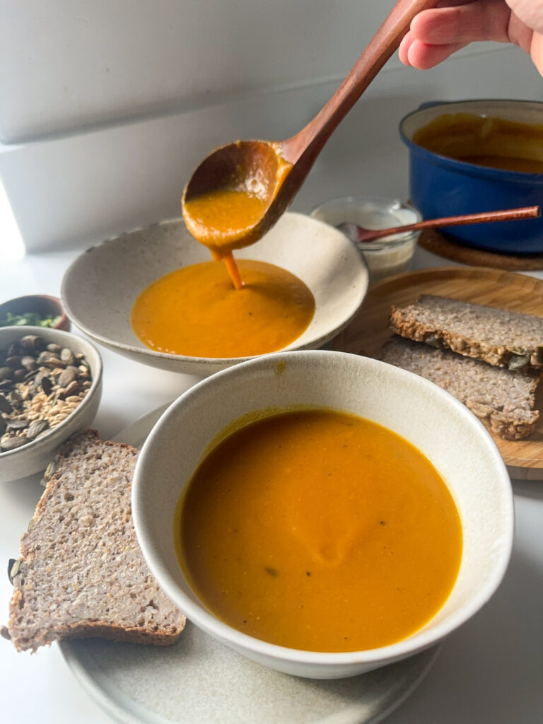 Pouring butternut squash soup into 2 bowls, surrounded by bread slices, seeds and herbs