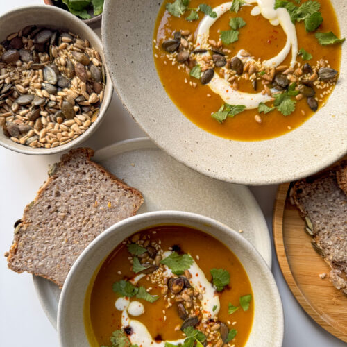 Roasted Squash Soup served in 2 bowls surrounded by bread, seeds and coriander