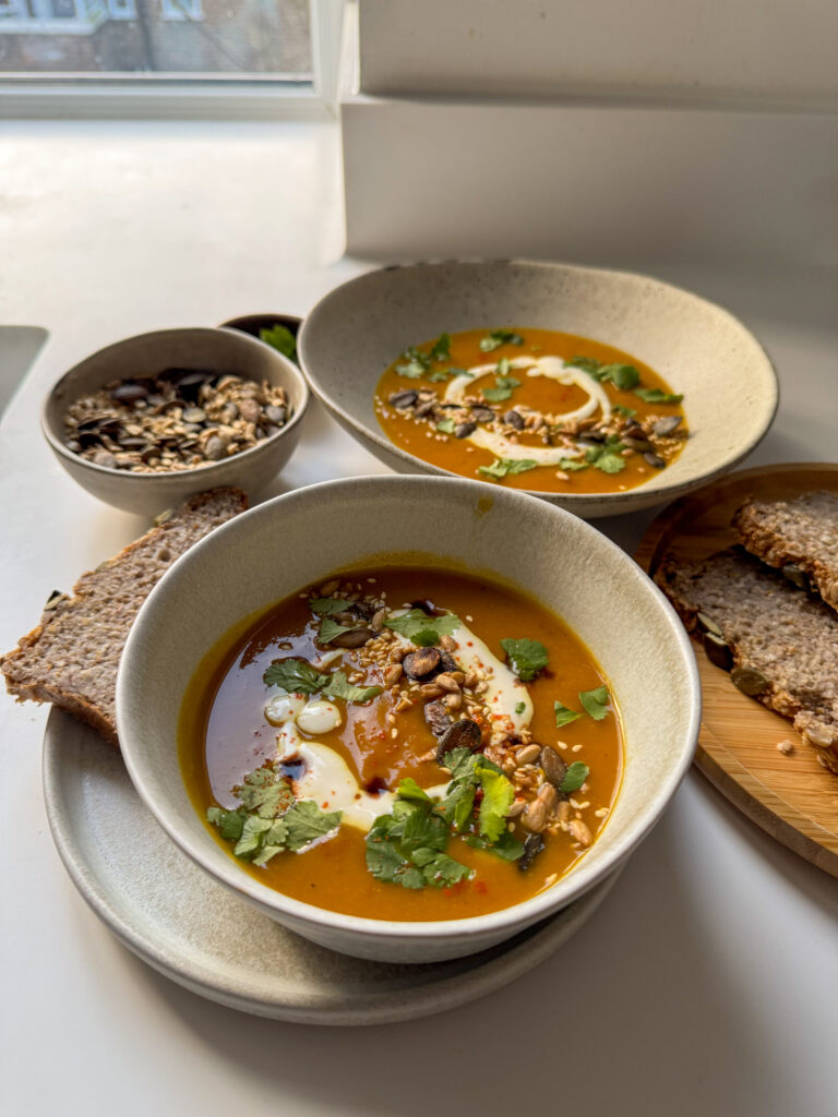 Roasted Squash Soup served in 2 bowls surrounded by bread, seeds and coriander