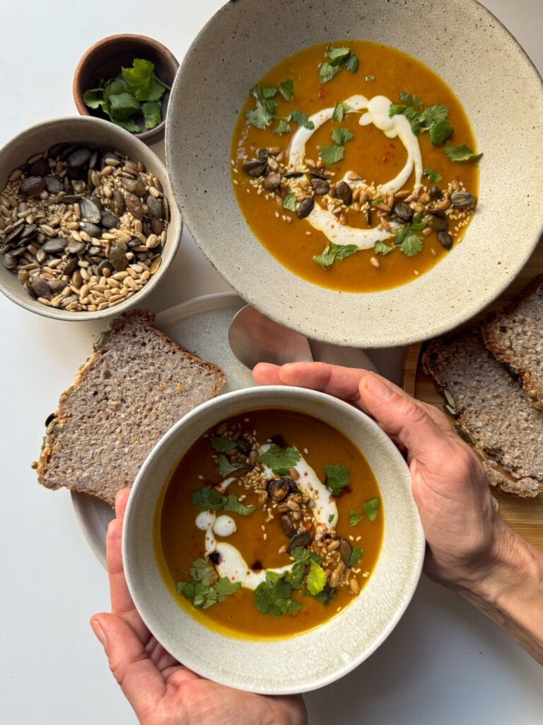 Roasted Squash Soup served in 2 bowls surrounded by bread, seeds and coriander